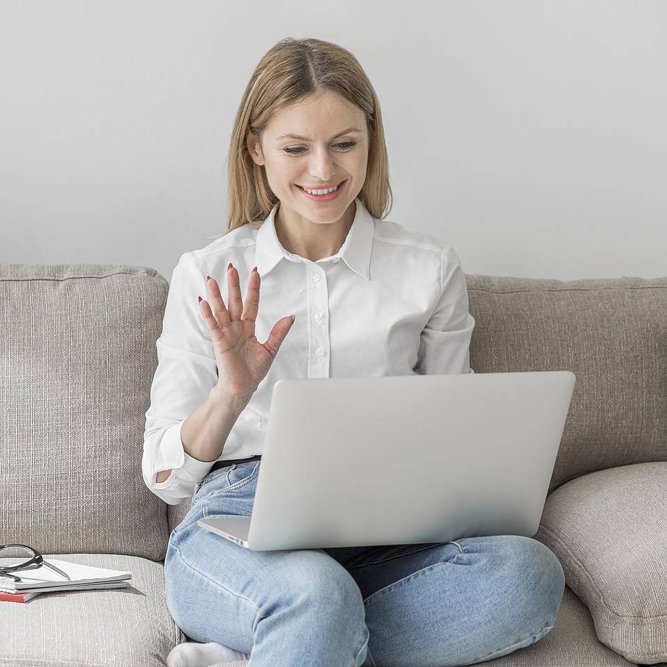 Eine Frau sitzt auf Zuhause auf ihrem Sofa und winkt lächelnd zum GPP-Online-Gruppe im Video-Call in ihrem Laptop, den sie auf ihren Knien balanciert.
